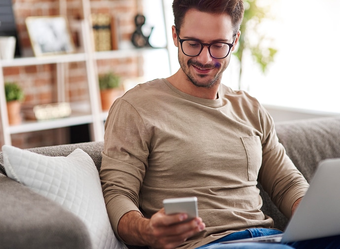 Homem sentado no sofá, com um notebook no colo, olhando para o telefone.